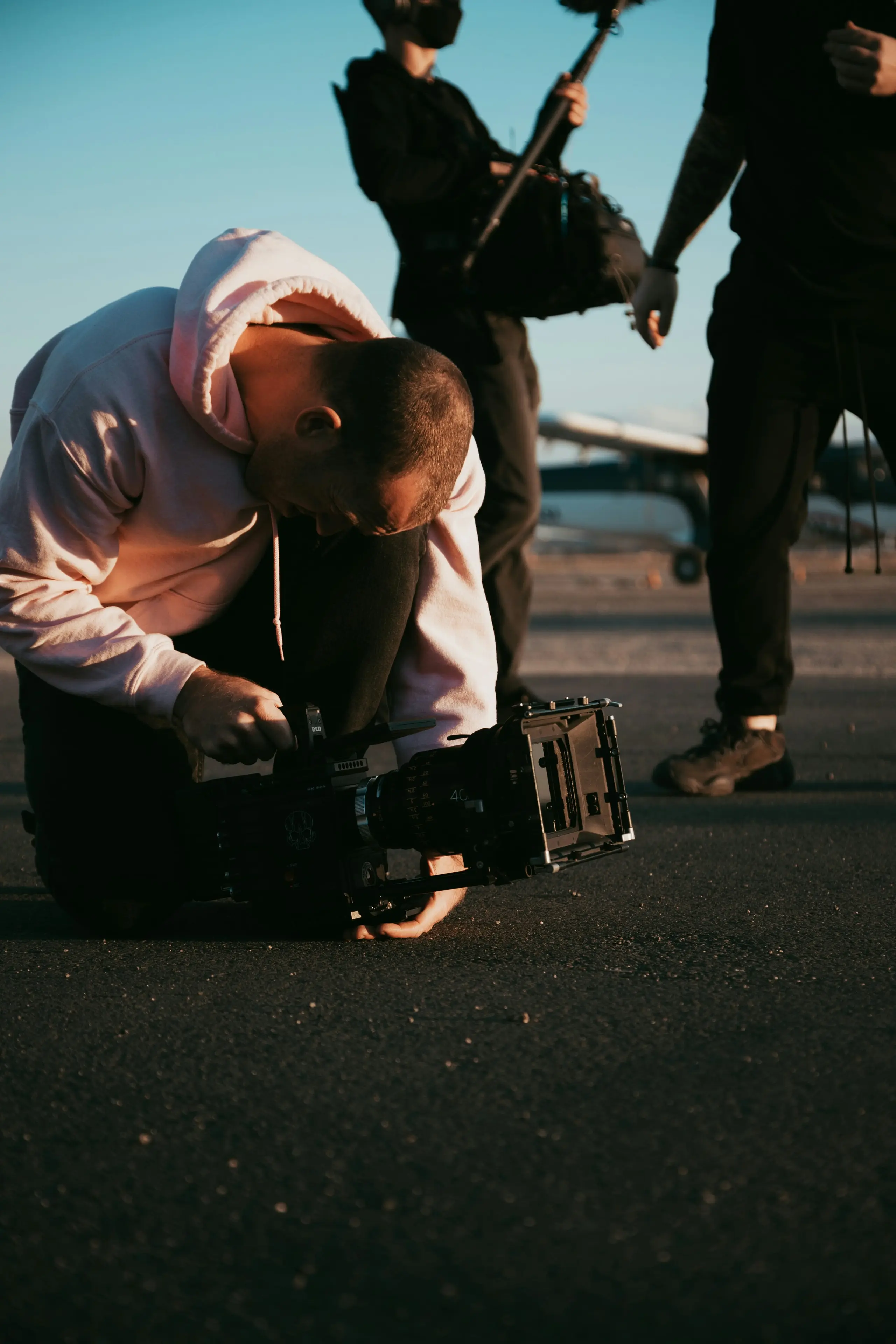 Crew member in a white hoodie crouched over a cinema camera on an airfield at golden hour