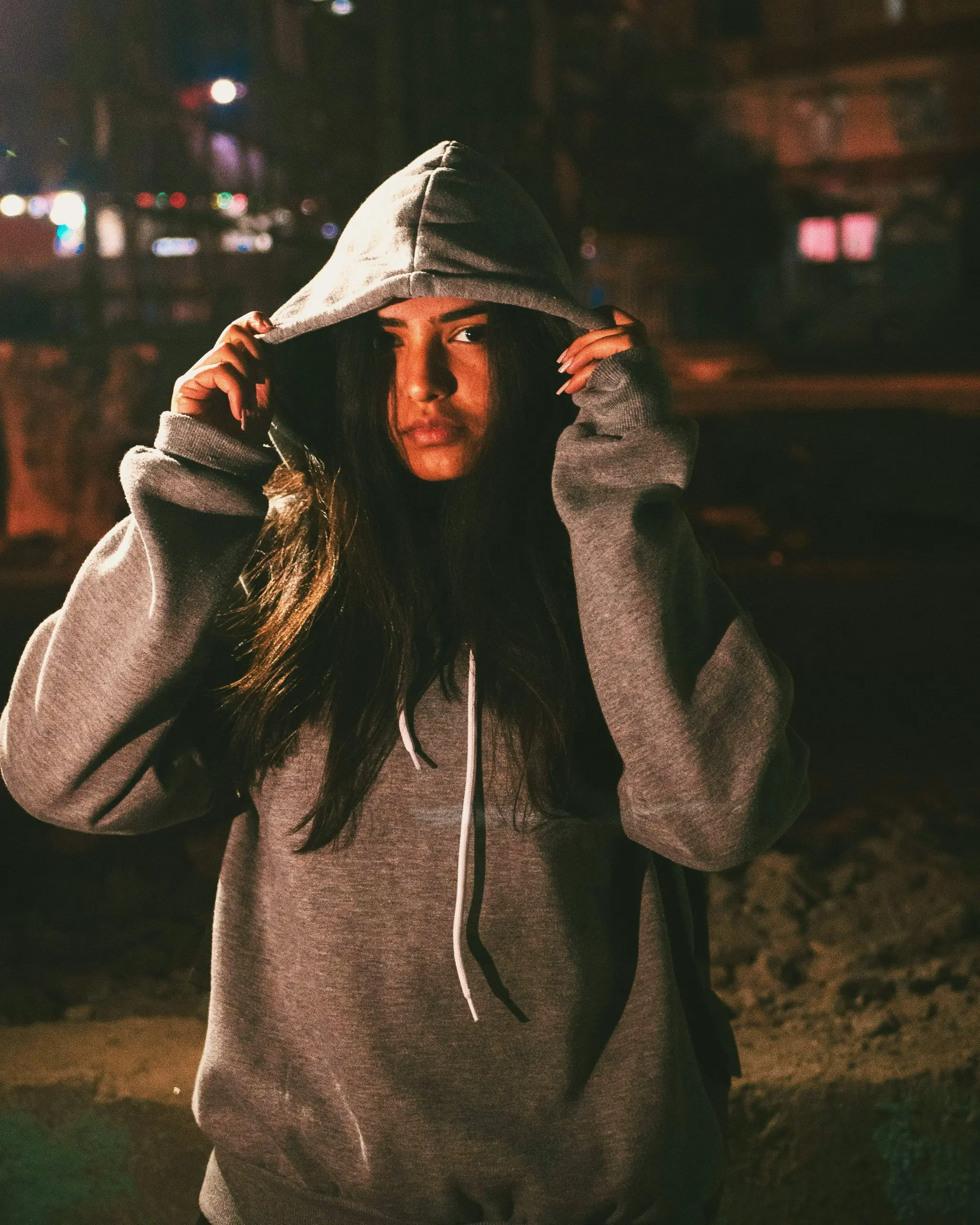 Grey hoodie, hood pulled up with both hands by a young woman at night, neutral gaze into the lens